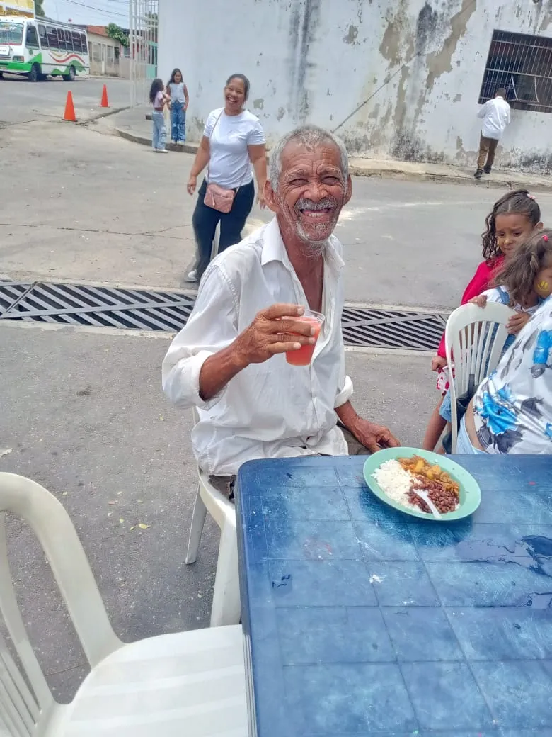 A man smiles and raises a cup during a shared meal at an outdoor table